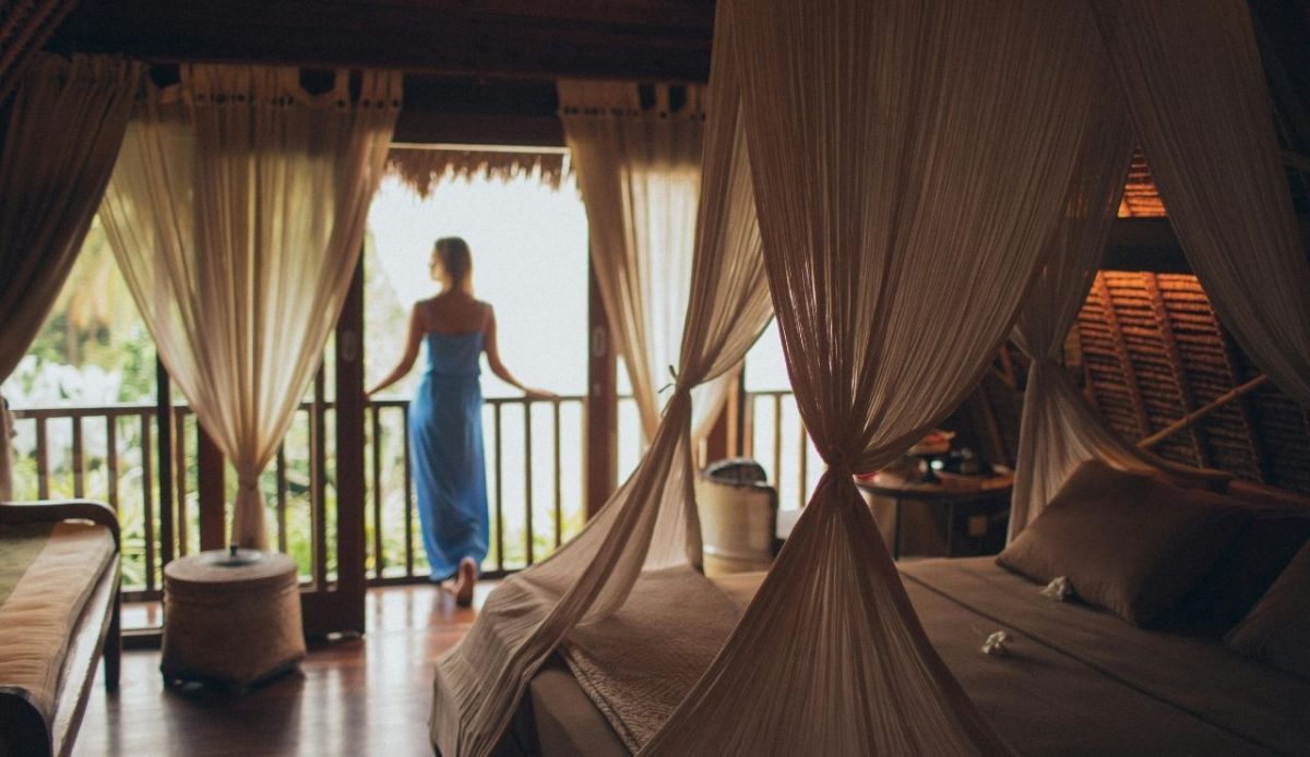Woman in a blue dress standing on a balcony of a tropical villa bedroom with canopy curtains
