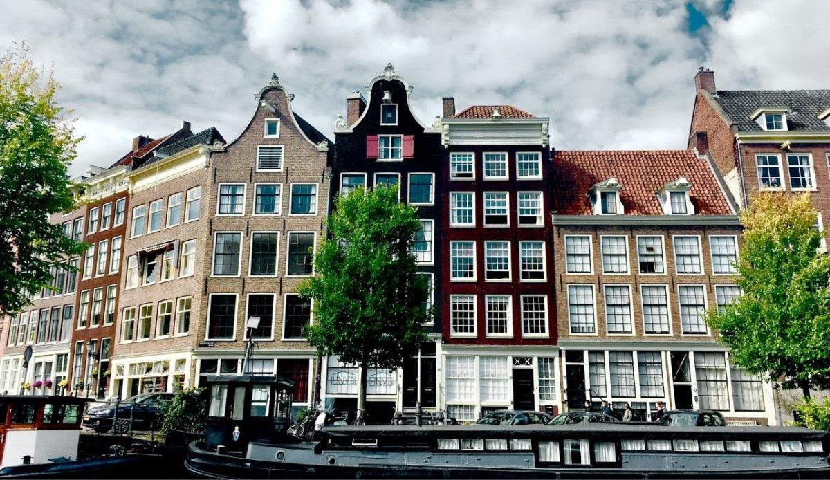 Traditional canal houses in Amsterdam with boats and trees in front

