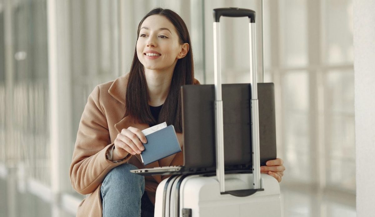 Smiling woman at the airport holding a passport and boarding pass while sitting beside her suitcase with a laptop placed on top
