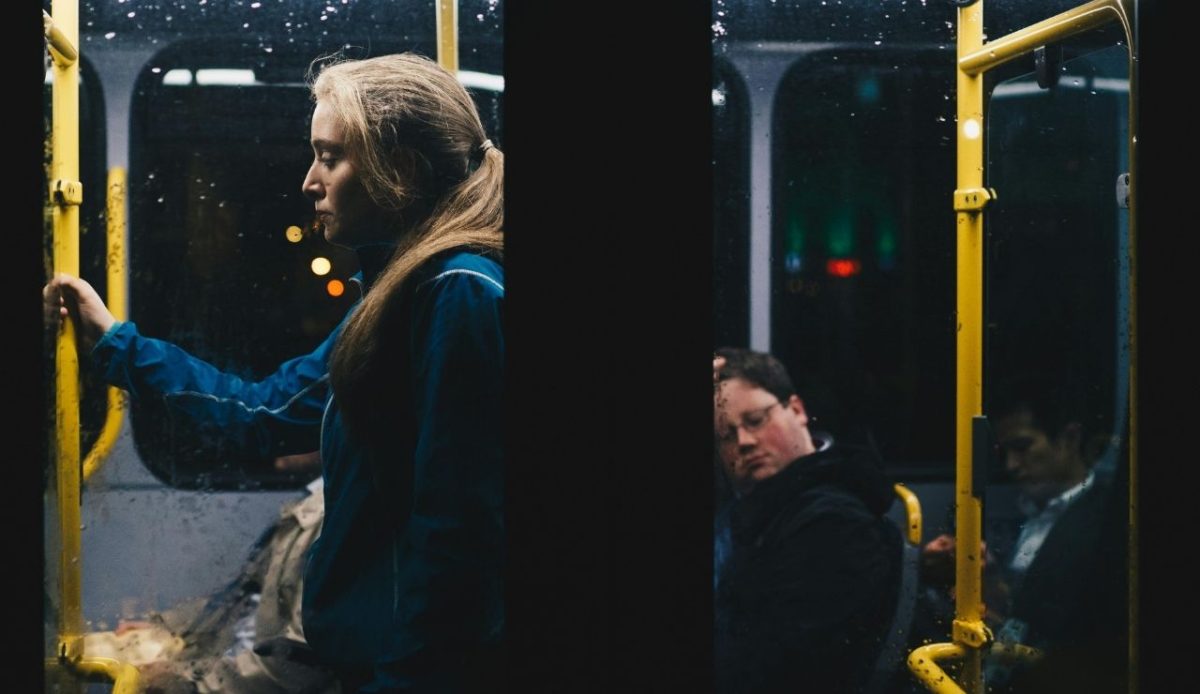 Young woman standing and holding a pole on a dimly lit bus at night while other passengers sit quietly
