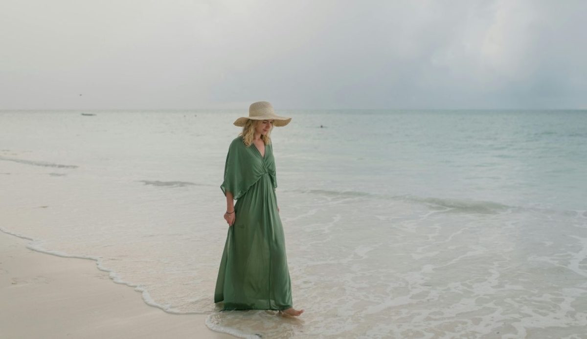 Woman in a green dress and wide-brimmed hat walking barefoot along the shoreline
