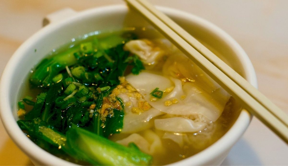 Bowl of wonton soup with green vegetables and chopsticks resting on the rim
