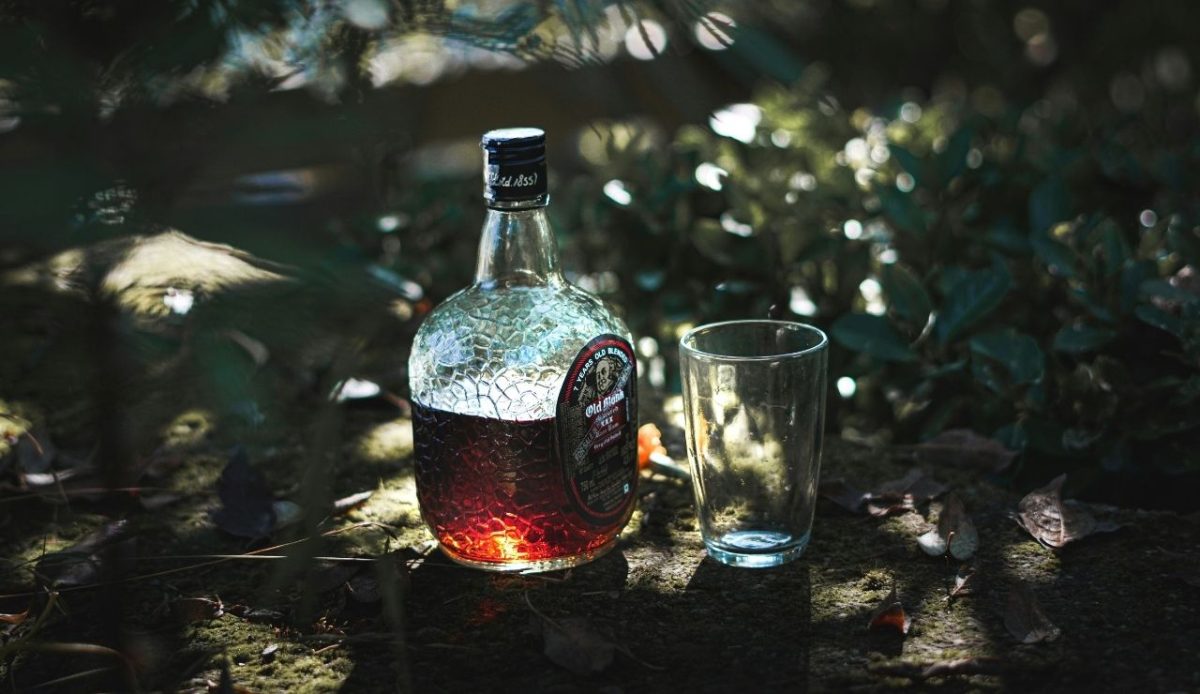 A glass bottle of dark liquor labeled "Old Monk" placed on the ground outdoors beside an empty glass, with leaves and greenery in the background.
