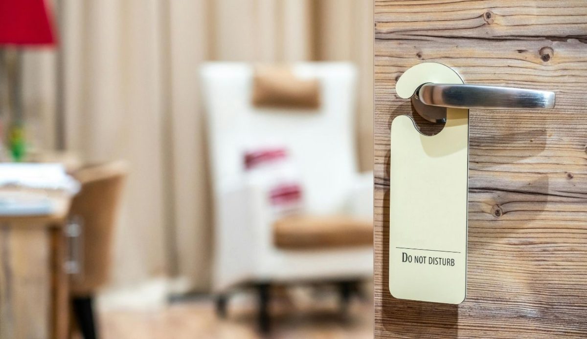 Close-up of a wooden hotel door with a silver handle and a "Do Not Disturb" sign hanging on it, with a blurred cozy room interior in the background
