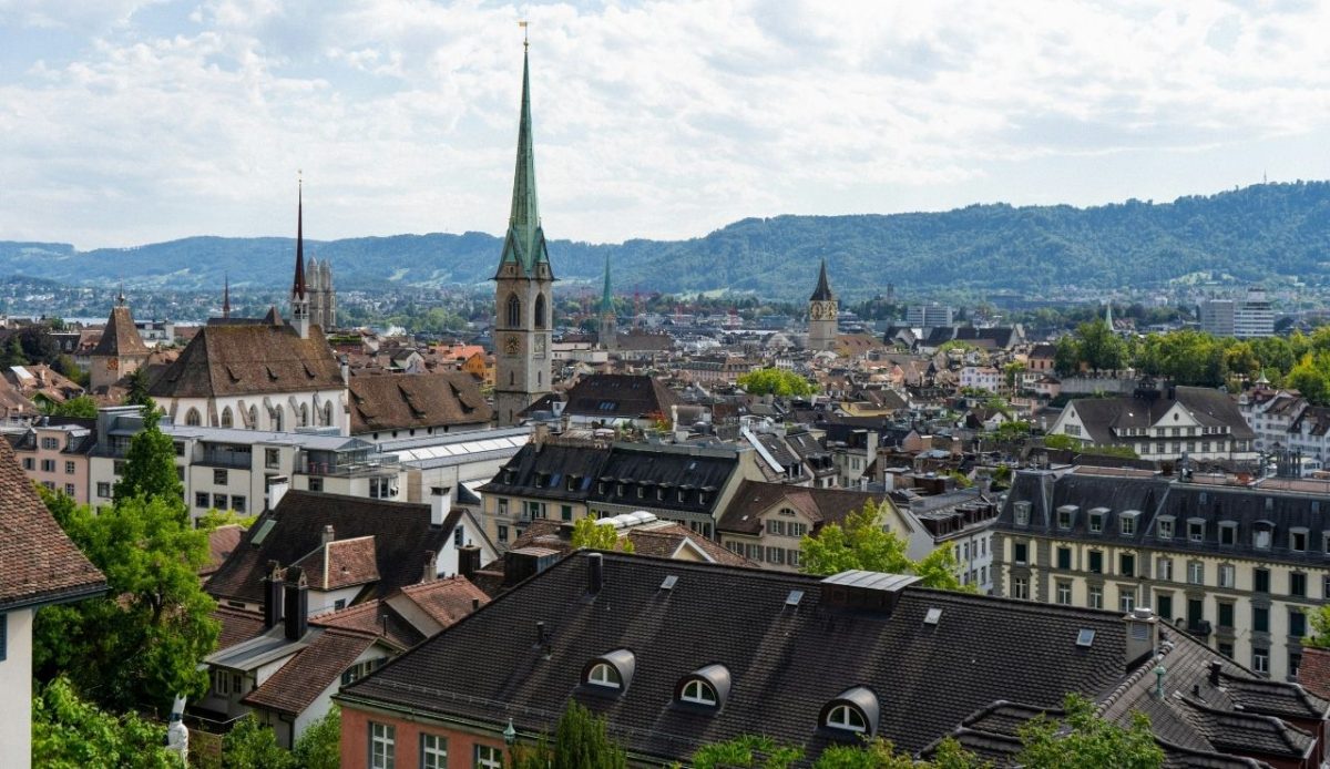 Panoramic view of Zurich with historic church towers and surrounding rooftops against a backdrop of green hills
