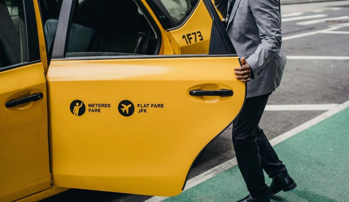Yellow taxi cab with open door showing metered fare and flat fare to JFK Airport, with a person in a gray suit entering
