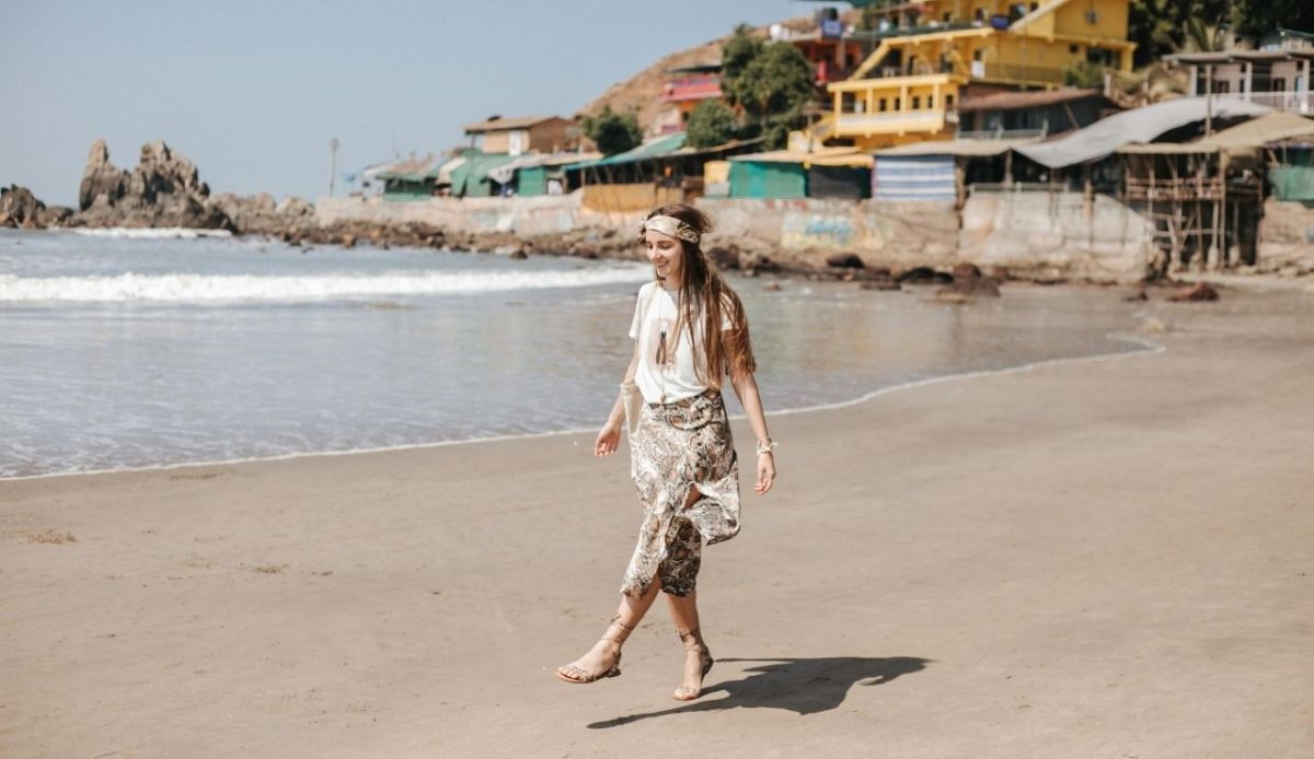 A woman in a bohemian outfit with a headband and patterned pants walking barefoot along a sandy beach, with colorful seaside buildings and waves in the background
