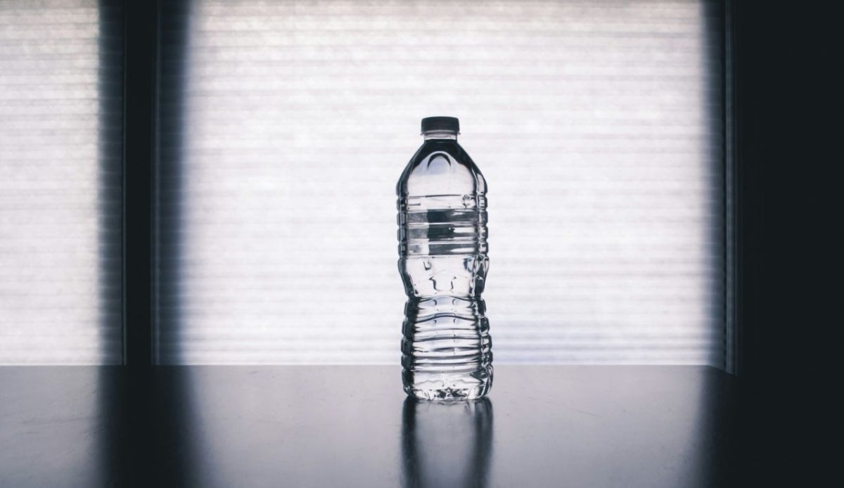 Clear plastic water bottle with a black cap placed on a reflective dark surface in front of a bright window.
