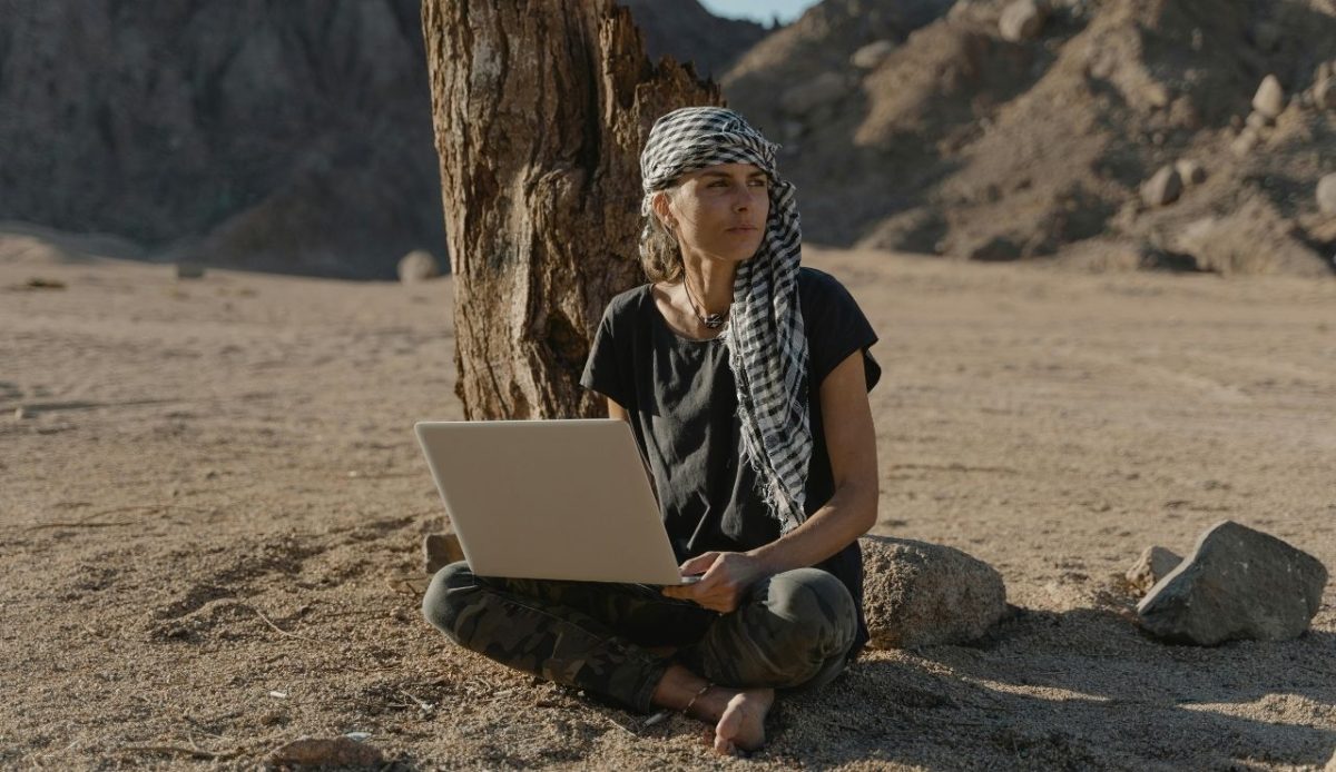 A woman wearing a headscarf and dark clothes sitting barefoot on desert sand under a tree while working on a laptop with rocky mountains in the background
