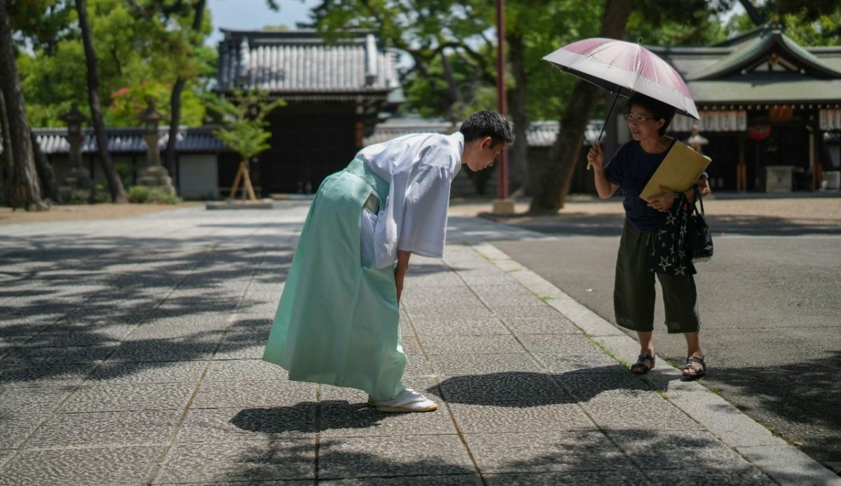 Man in traditional Japanese attire bowing to a woman with an umbrella in a temple courtyard
