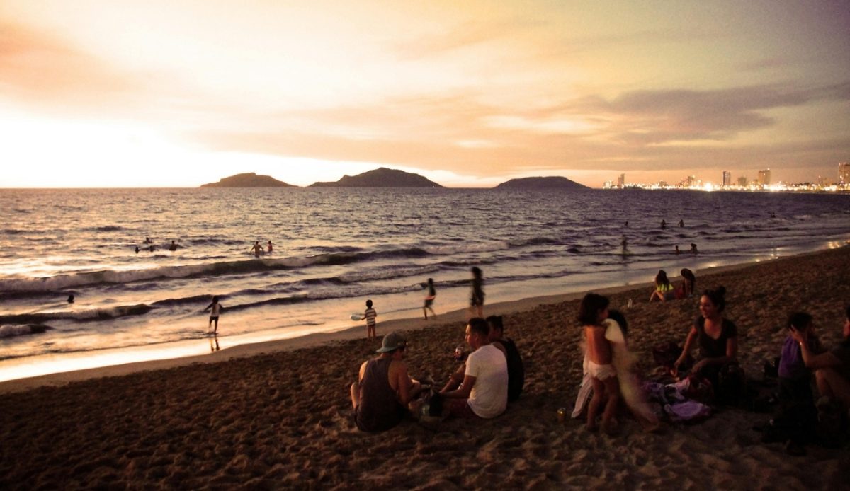 People relaxing on the beach at sunset, with the ocean and hills in the background, likely in Mazatlán, Mexico 