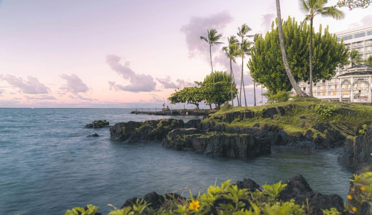 9 Best Hawaiian Islands to Visit for Every Kind of Traveler 7 Coastal view of Hilo Bay, Hawaii, with palm trees, volcanic rocks, and calm ocean water at sunset