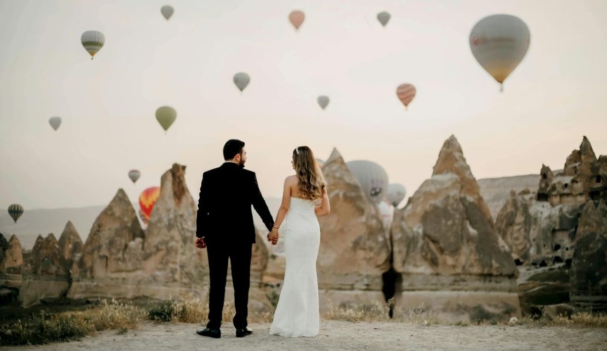 Back View of Groom and Bride with Tuff Rock Formations and Hot Air Balloons in Sky, Göreme, Turkey                    