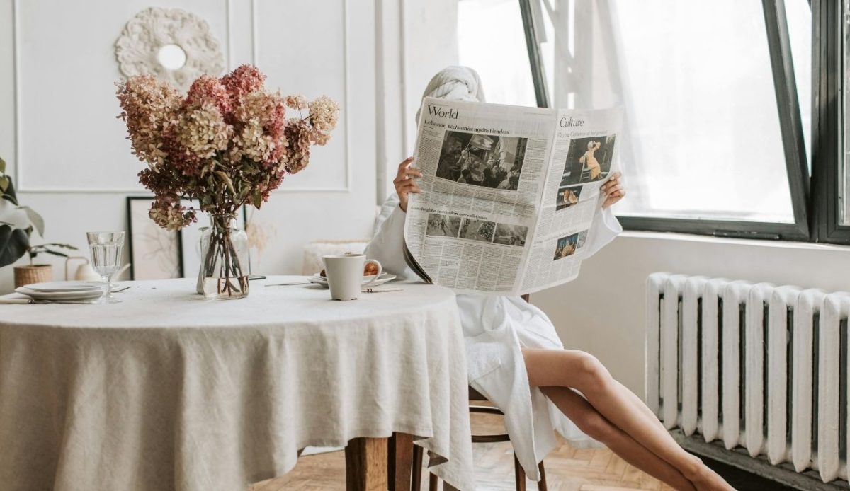 Person in White Robe Sitting on Brown Chair while Holding a Newspaper  in hotel room      