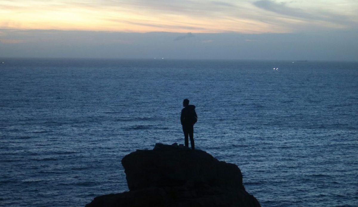 Silhouette of a person standing on a rocky cliff overlooking the ocean at dusk                    