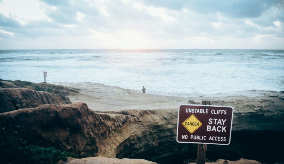 Stay back sign over cliffs and as man looks over a stormy sea                        