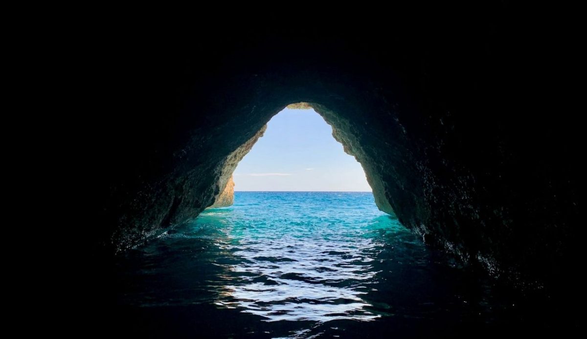 View from inside a sea cave opening onto turquoise water and open ocean under a clear sky                    