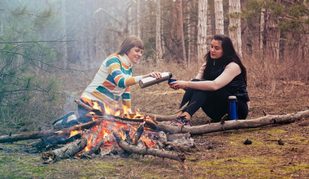 Two women camping in the forest, pouring hot drinks by a campfire 