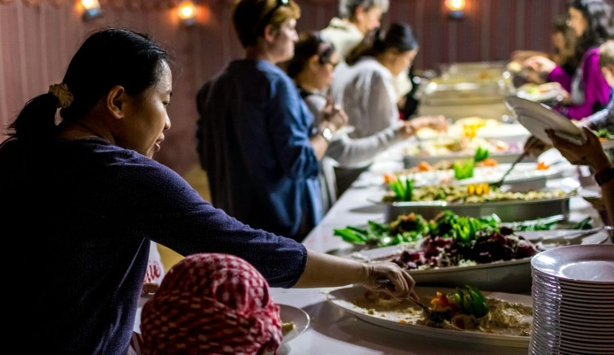 People standing close together at a busy buffet line, sharing serving utensils and leaning over open trays of food                    