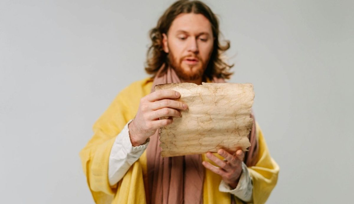 Person dressed in biblical-style robe holding and reading an aged parchment scroll against a plain background                             
