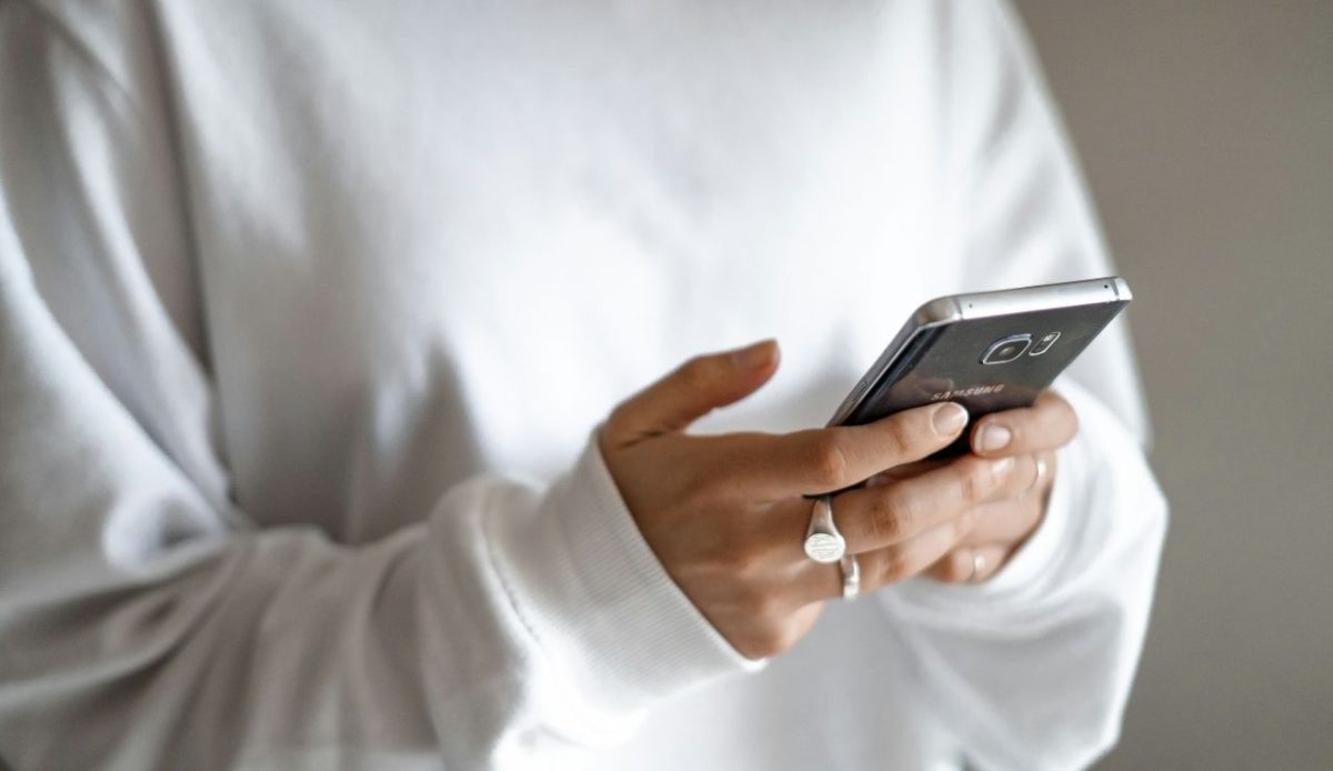 Person wearing a light sweatshirt holding a smartphone with both hands, viewing the screen indoors against a neutral background                              