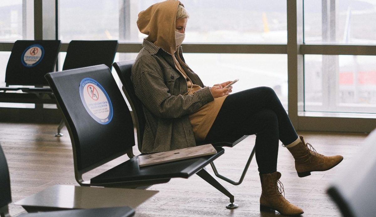 Traveler wearing a mask and hoodie using phone while waiting at airport gate 