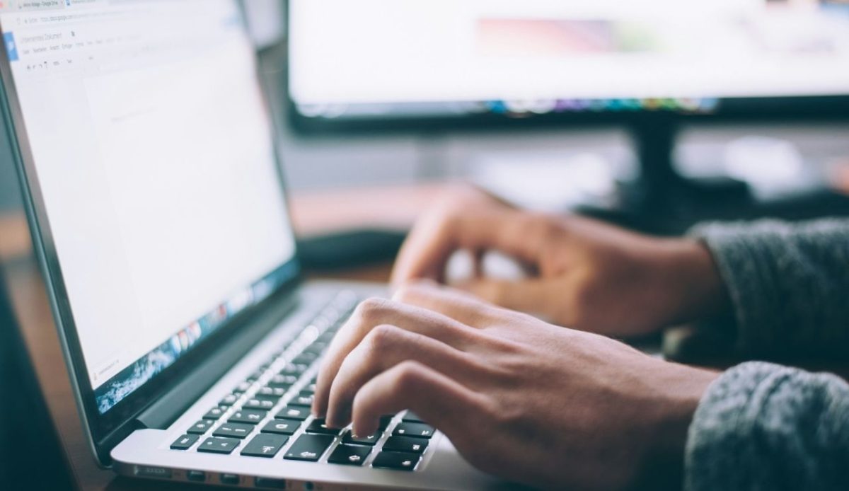 Close up of hands typing on a laptop keyboard with blurred computer screens in the background                             