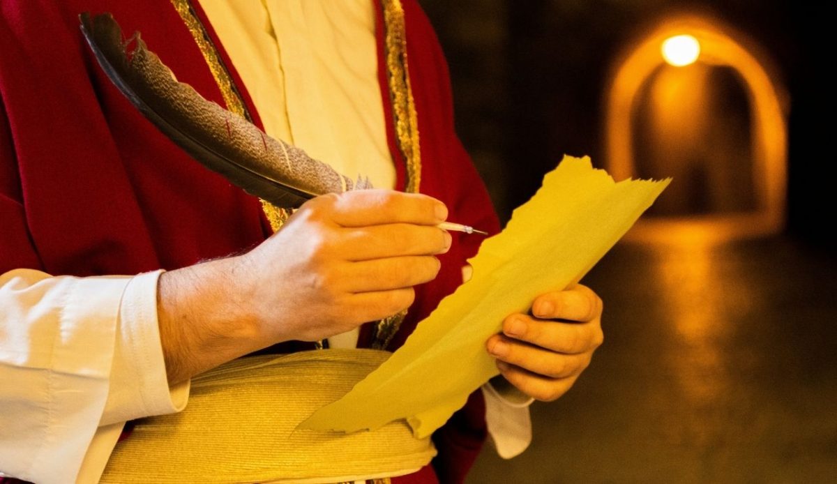Person in red ceremonial robe writing with a quill on yellow parchment inside a dimly lit historic interior with arched doorway in background                               