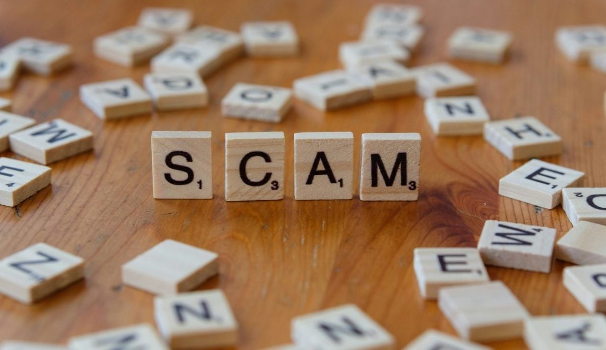 Wooden letter tiles spelling the word Scam on a table, surrounded by scattered tiles, symbolizing fraud targeting religious travelers                             
