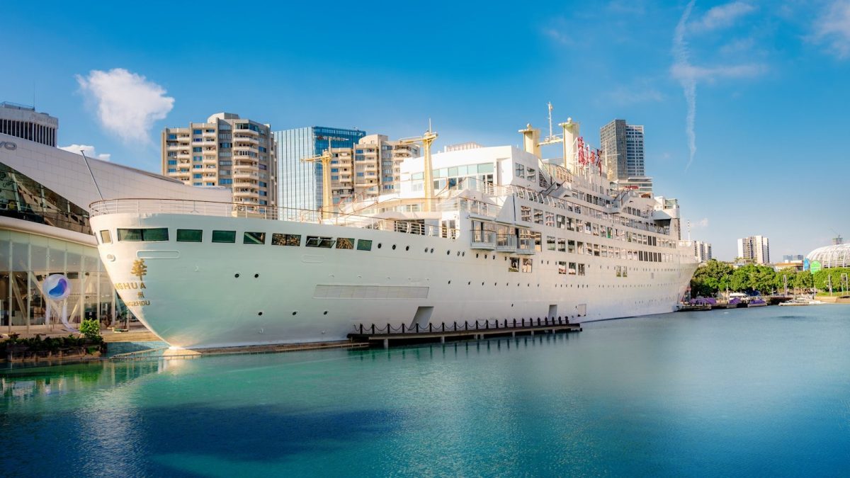 A large white cruise ship docked by a modern city.