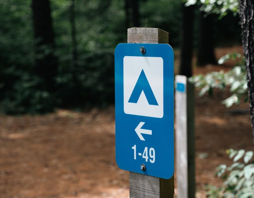 5 Campground First-Night Checks Travelers Usually Wish They Did Before Dark 4 Blue campsite direction sign in forest pointing to sites 1 49, surrounded by trees and nature.