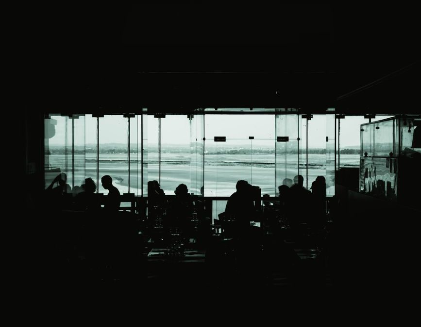 Silhouetted people in an airport lounge against large windows overlooking the runway.
