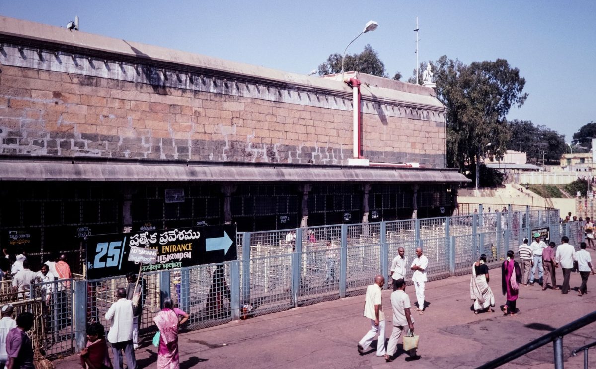 People entering a temple in India on a sunny day. Entrance sign and local architecture are visible.