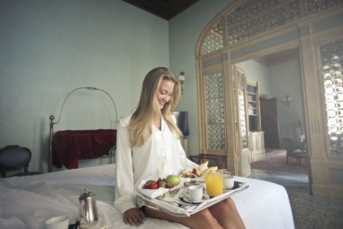 Happy young blonde sitting on bed in luxury hotel room having tray with served delicious breakfast on knees enjoying exotic resort