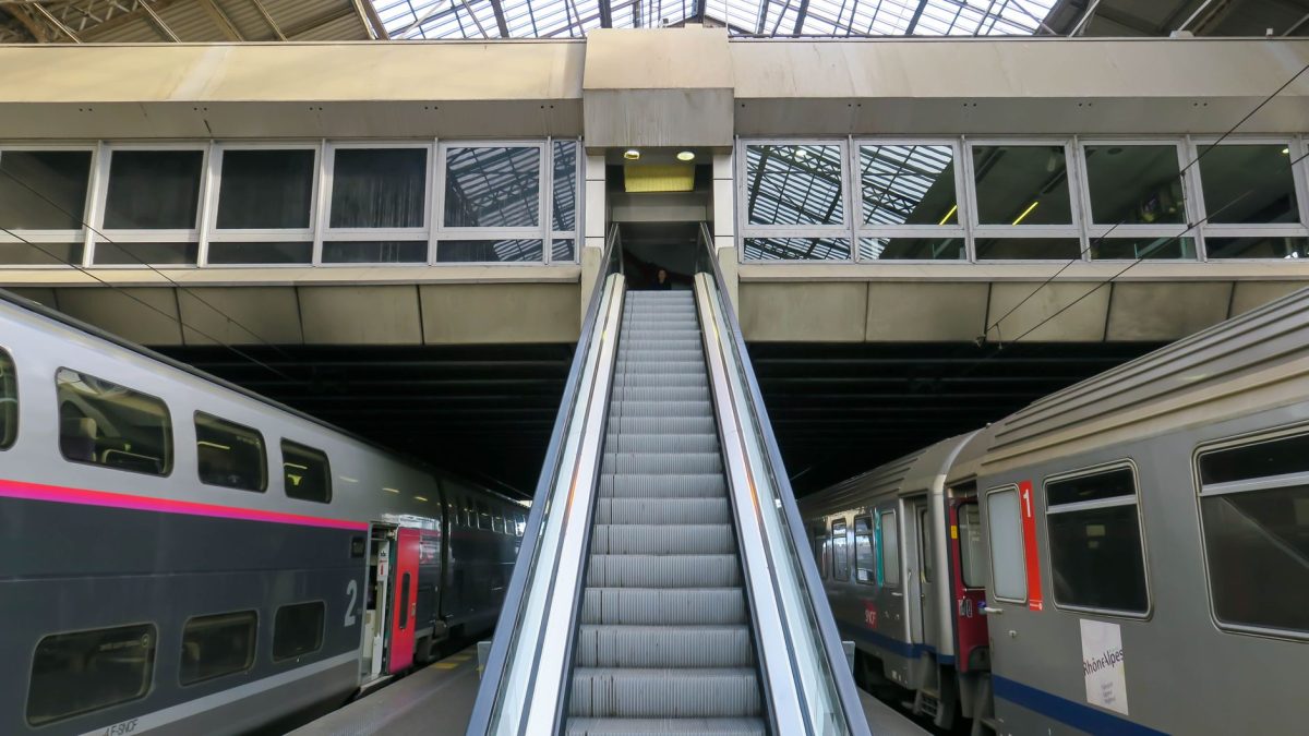 View of an escalator leading to platforms in a modern train station with trains on either side.