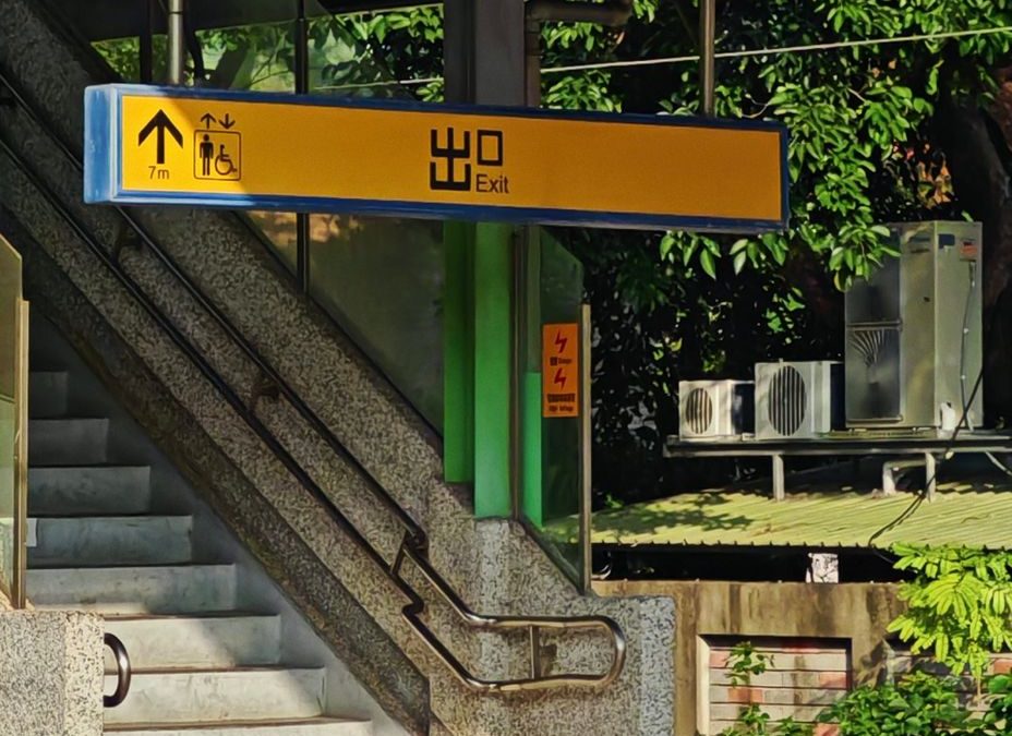 Staircase and exit sign at a sunny train station, featuring greenery and urban architecture.