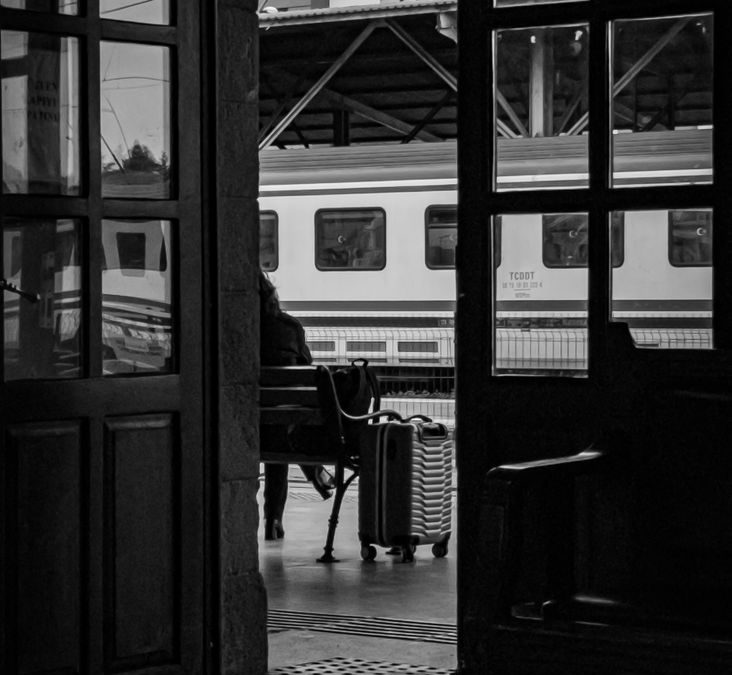 A person waits at a train station with a suitcase, framed by a doorway, in black and white.