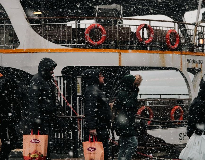 People walking by a ferry during snowfall, carrying shopping bags in a bustling urban setting.