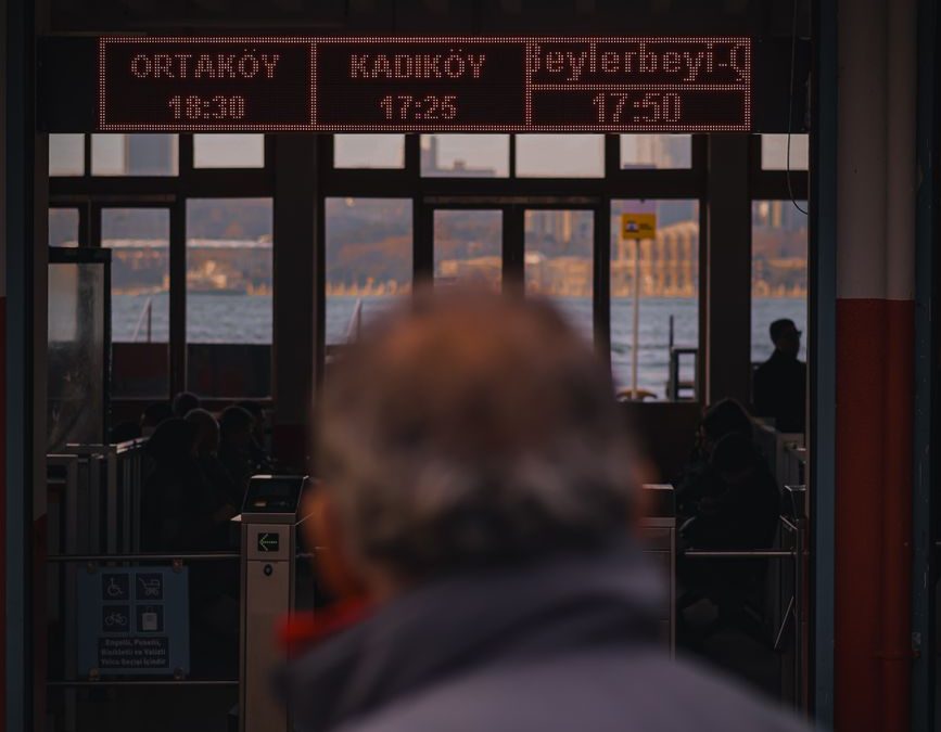 A person stands at a ferry terminal, looking at a timetable for Ortaköy, Kadiköy, Beylerbeyi routes.