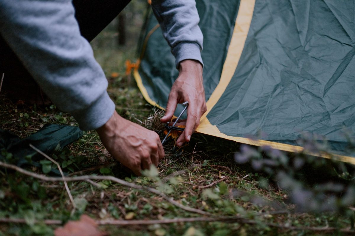 A close up of a person securing a tent peg in a forested campsite, depicting outdoor camping activities.