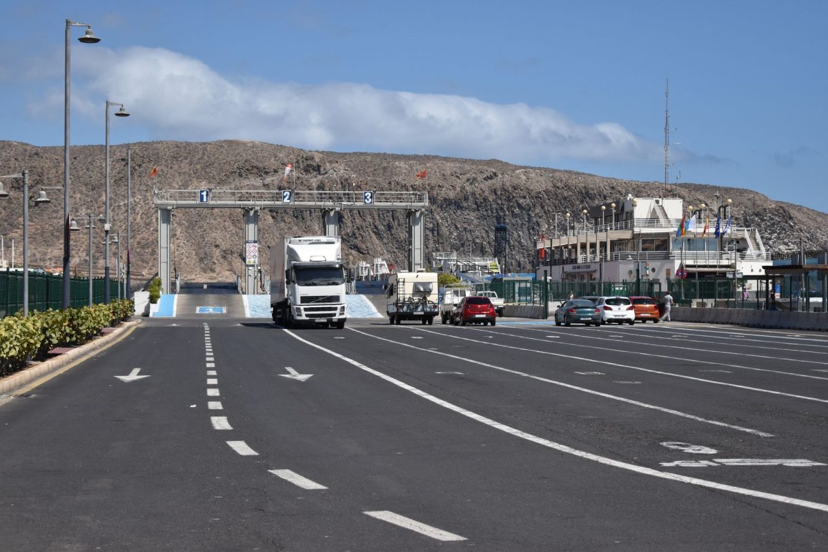 A ferry terminal with trucks and cars under a clear blue sky and dry hills in the background.