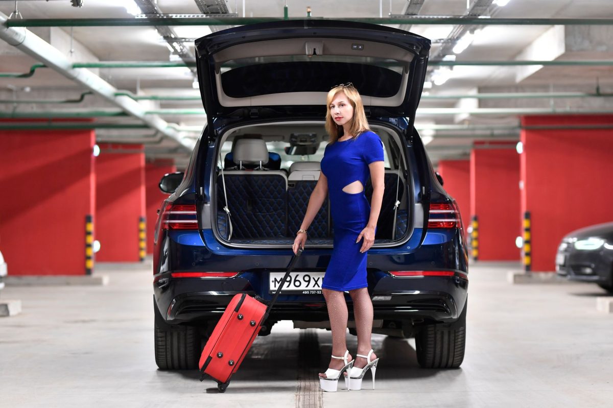 Stylish woman in blue dress posing with luggage by a car in a parking garage.
