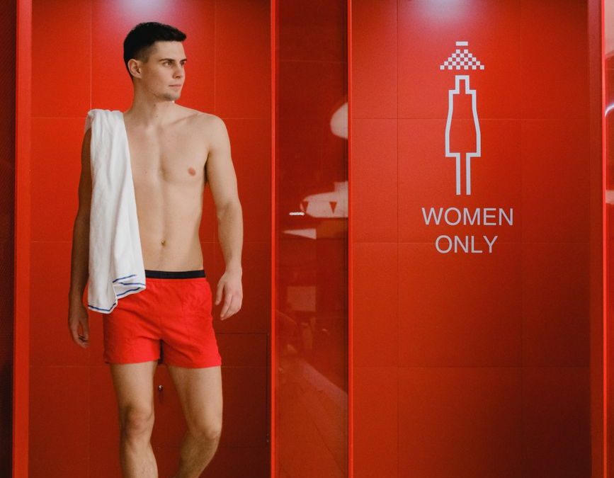 A man in red swim trunks exits a bright red shower room with a &amp;#39;Women Only&amp;#39; sign.