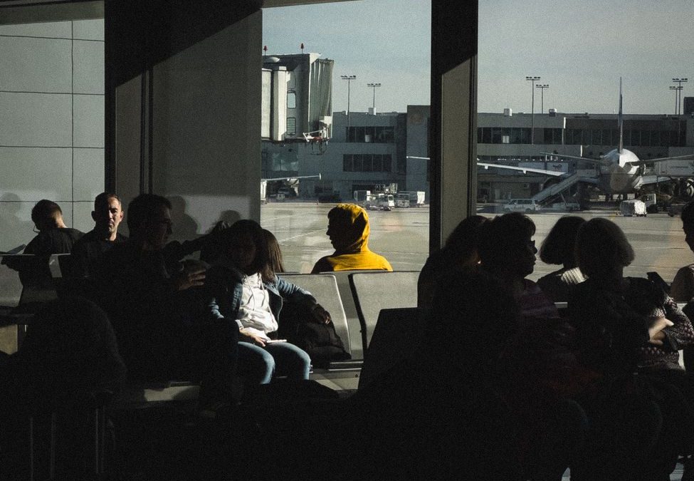 Silhouetted travelers in an airport terminal waiting area near large windows with outside tarmac view.