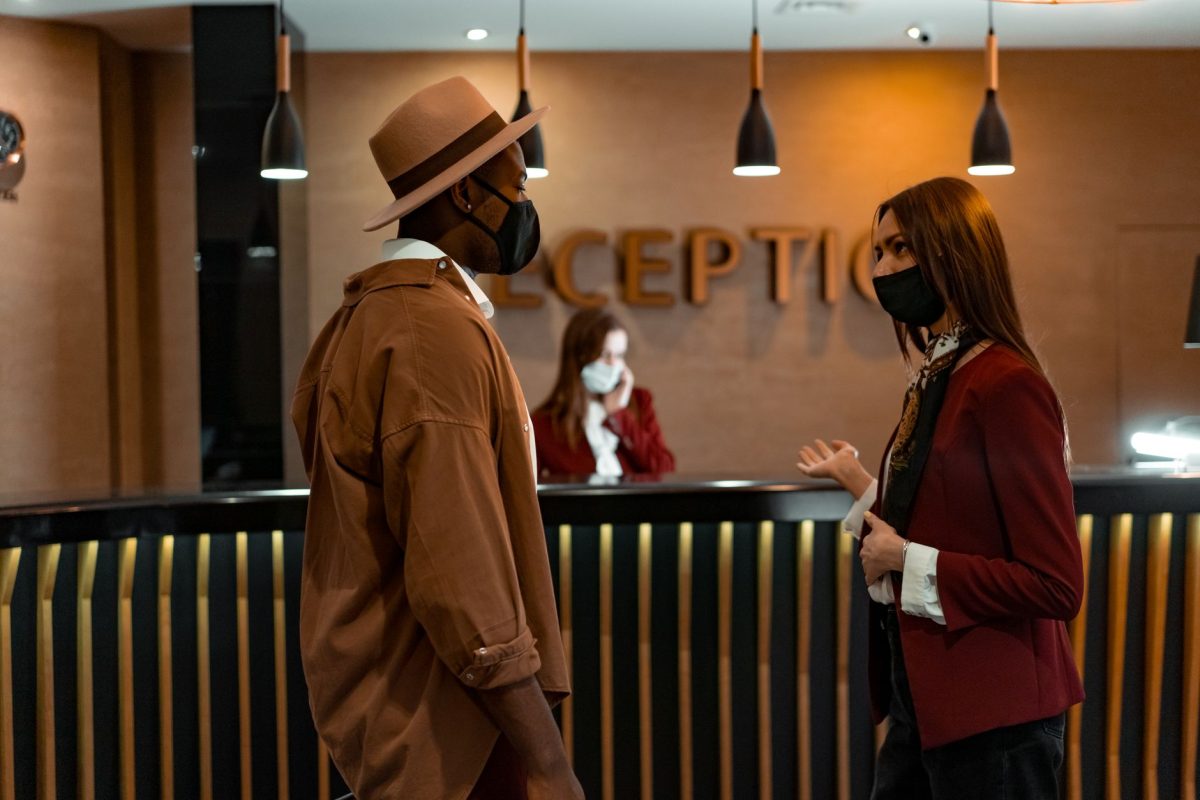 Two people in face masks interacting at a hotel reception desk during the COVID 19 pandemic.