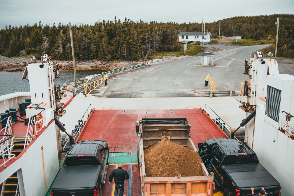 Trucks disembarking a ferry at a forested coastal location on a cloudy day.