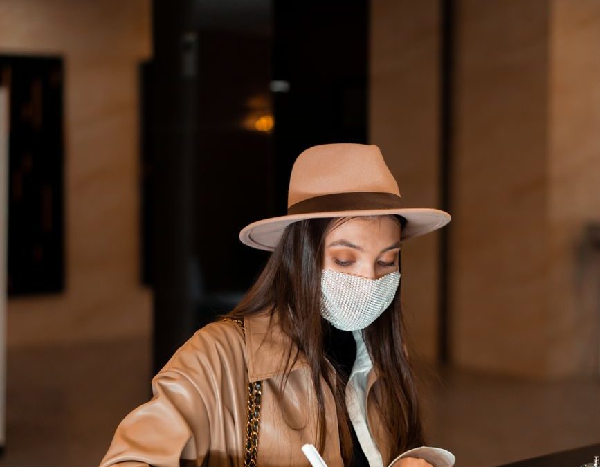 A woman wearing a face mask and stylish hat checks into a hotel at the reception desk.