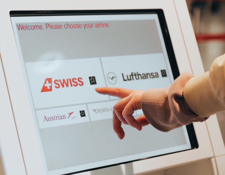 5 Bag-Drop Slipups That Can Derail a Travel Morning 2 Hand interacting with airport kiosk for airline selection. Modern travel technology.