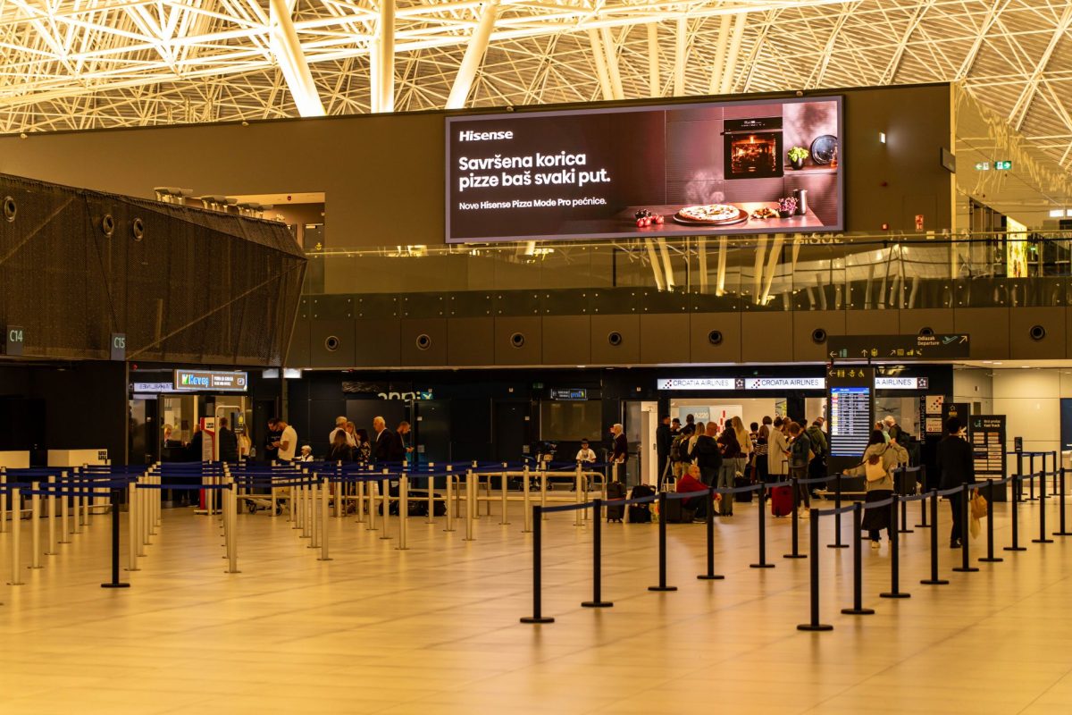 5 Bag-Drop Slipups That Can Derail a Travel Morning 5 Travelers check in at Zagreb Airport with large advertising screen above.
