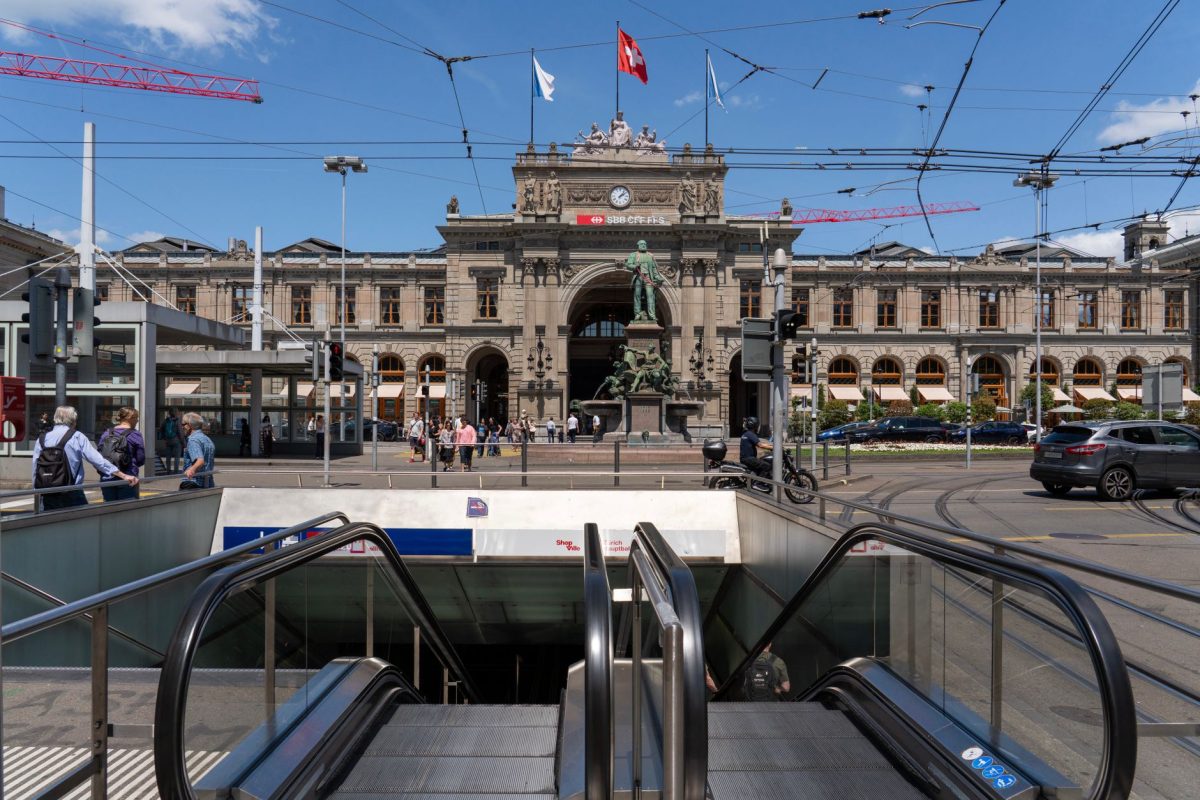 View of Zurich Hauptbahnhof facade with people and cars under a clear blue sky.